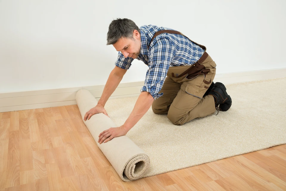 Un homme en chemise à carreaux et salopette beige déroule un tapis beige dans une pièce aux murs blancs sur parquet bois clair