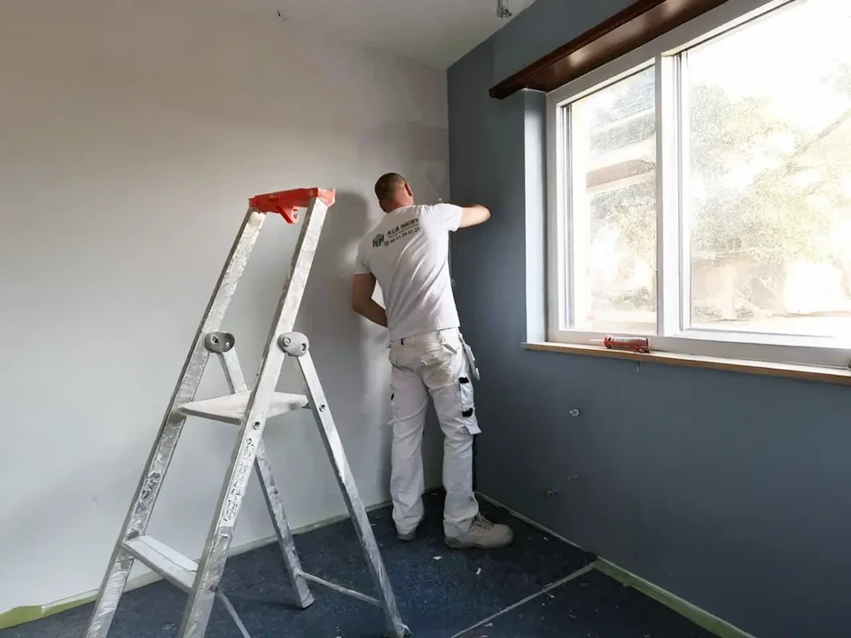 Deux ouvriers en combinaisons bleue et verte travaillent à genoux sur un chantier de rénovation intérieure, manipulant des outils de mesure près d'un mur blanc